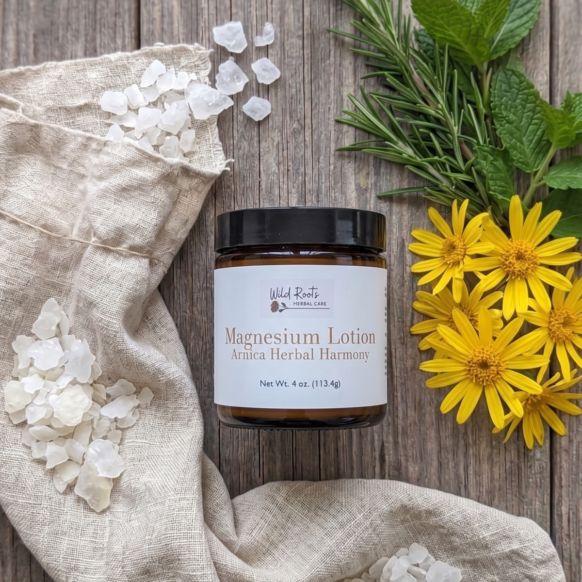 A jar of Wild Roots Organic Magnesium Lotion sits on a rustic wooden surface, styled with a beige linen bag, white bath salts, rosemary, mint, and yellow flowers.