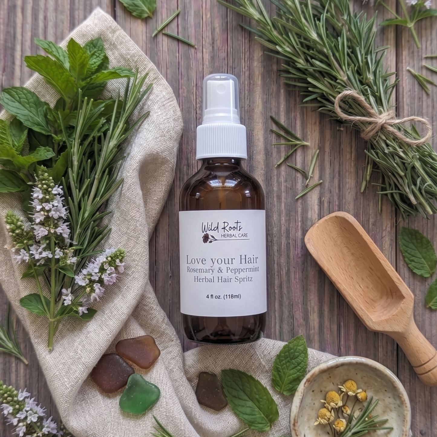 A bottle of Wild Earth Hair Savior hair spray with rosemary and peppermint ingredients, styled in a flat lay on a rustic wooden table with fresh rosemary, mint leaves, a wooden scoop, and a small bowl of dried herbs.