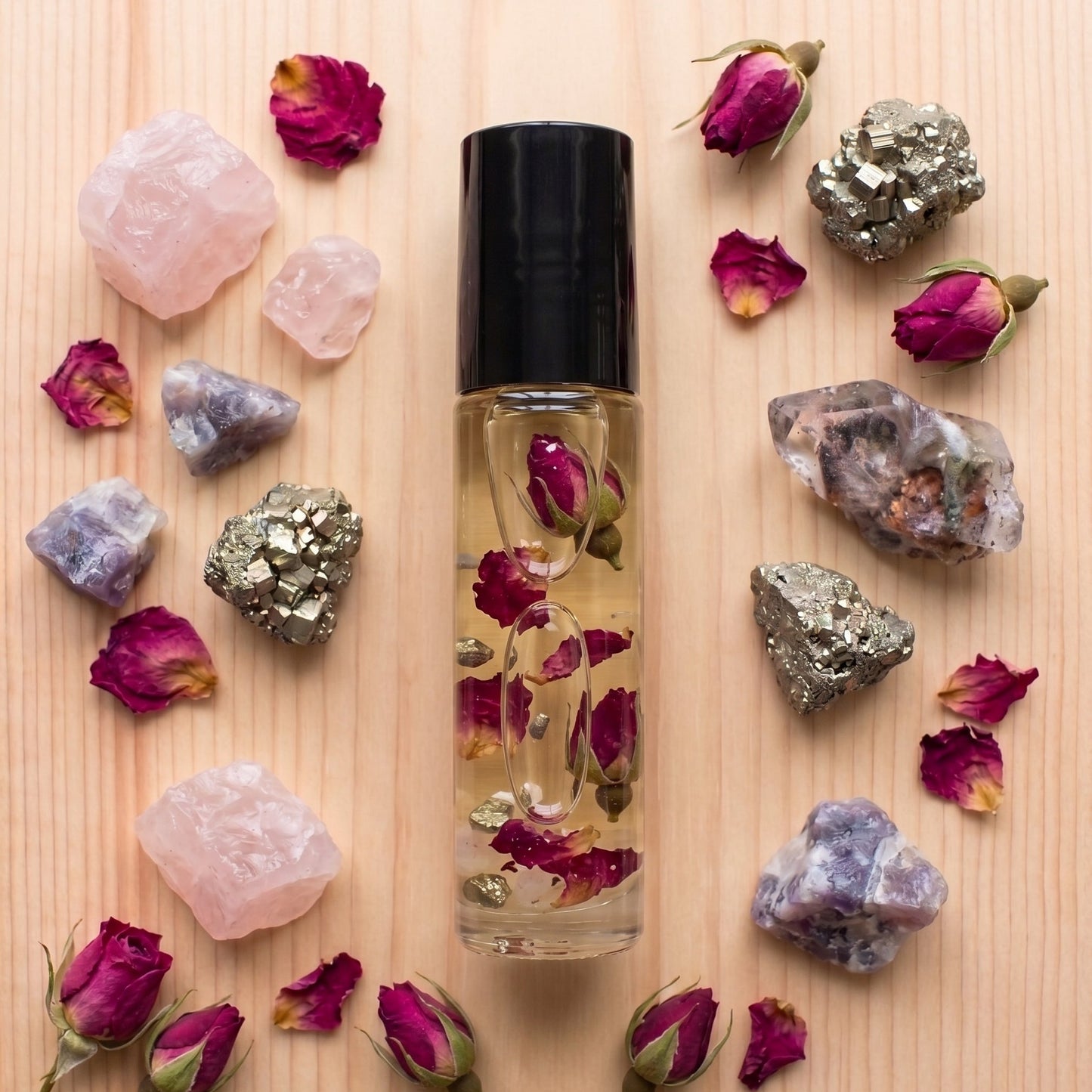 A clear glass roller bottle with a black cap, filled with a golden liquid and dried rosebuds, arranged on a light wood surface and surrounded by pink, purple, and silver crystals with scattered rose petals.