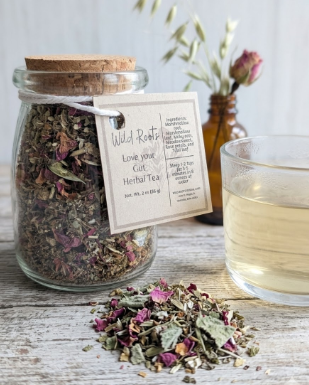 Glass jar of dried herbs with a label, surrounded by loose herbs and a glass of tea on a wooden surface.