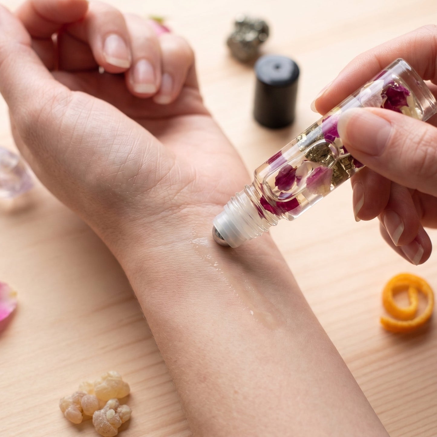 A close-up of a person's forearm and hand receiving a roll-on oil application from a clear glass roller bottle containing dried flowers, set on a light wooden surface with small decorative crystals and petals.