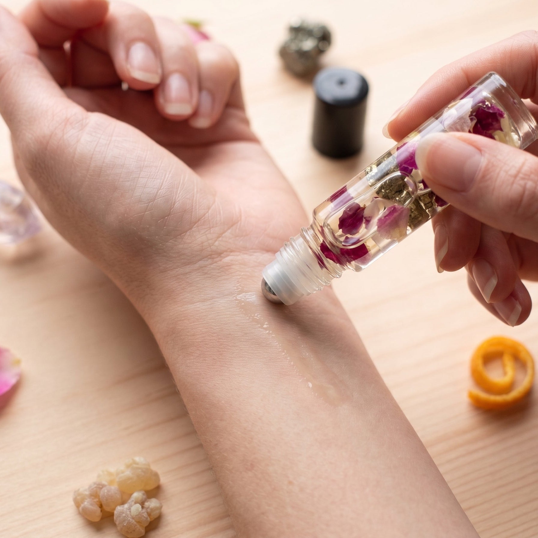 A close-up of a person's forearm and hand receiving a roll-on oil application from a clear glass roller bottle containing dried flowers, set on a light wooden surface with small decorative crystals and petals.