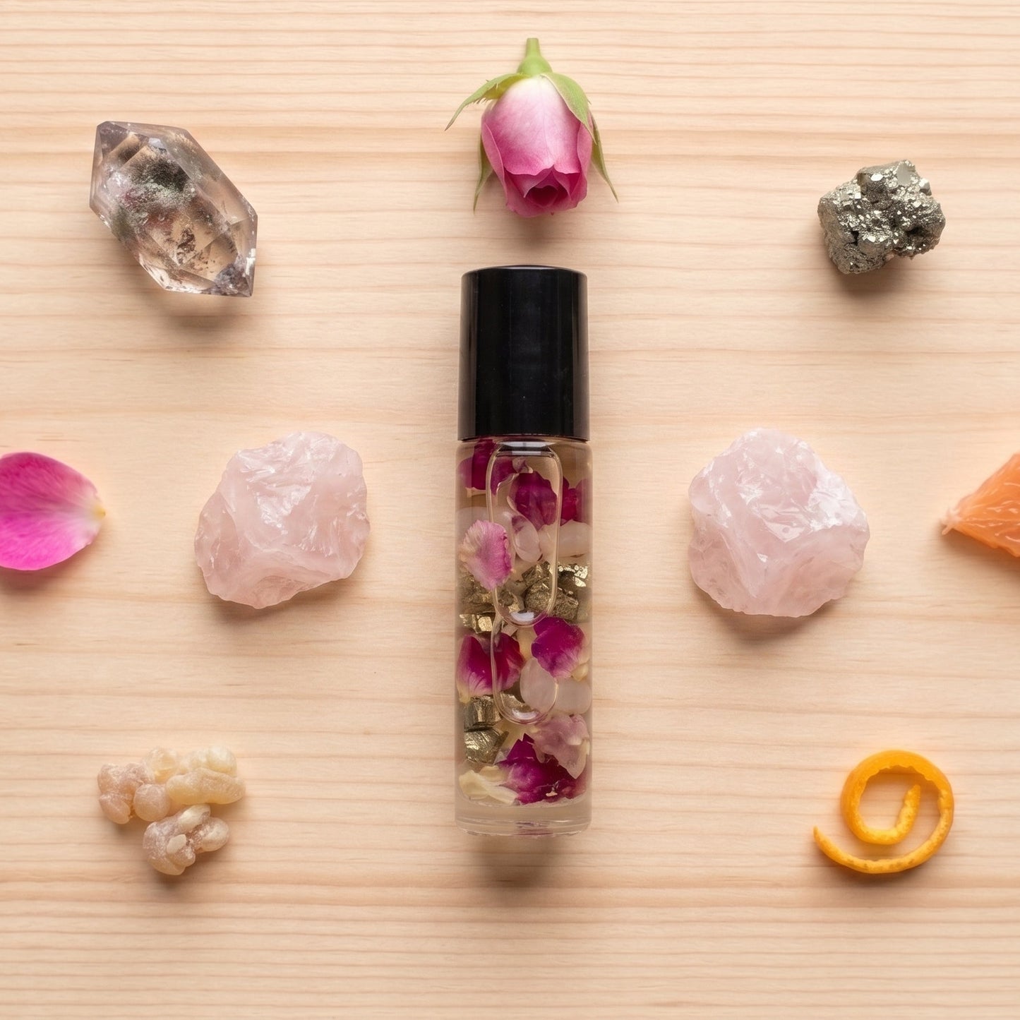 A clear glass roller bottle with a black cap, filled with dried rose petals and gold flakes, arranged on a light wood background with pink crystals, rose petals, a rose bud, a crystal cluster, a piece of orange, and a curled orange peel.