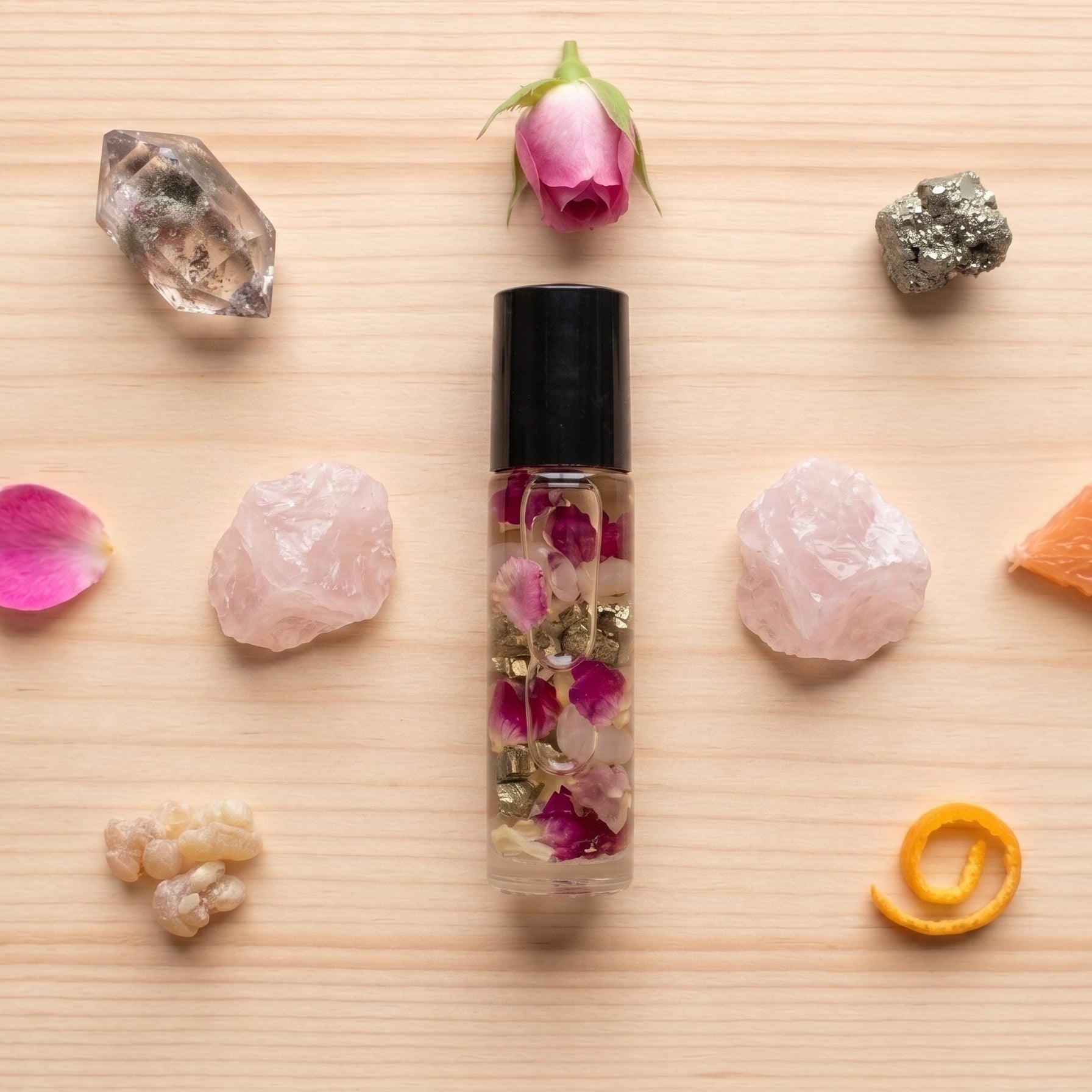 A clear glass roller bottle with a black cap, filled with dried rose petals and gold flakes, arranged on a light wood background with pink crystals, rose petals, a rose bud, a crystal cluster, a piece of orange, and a curled orange peel.