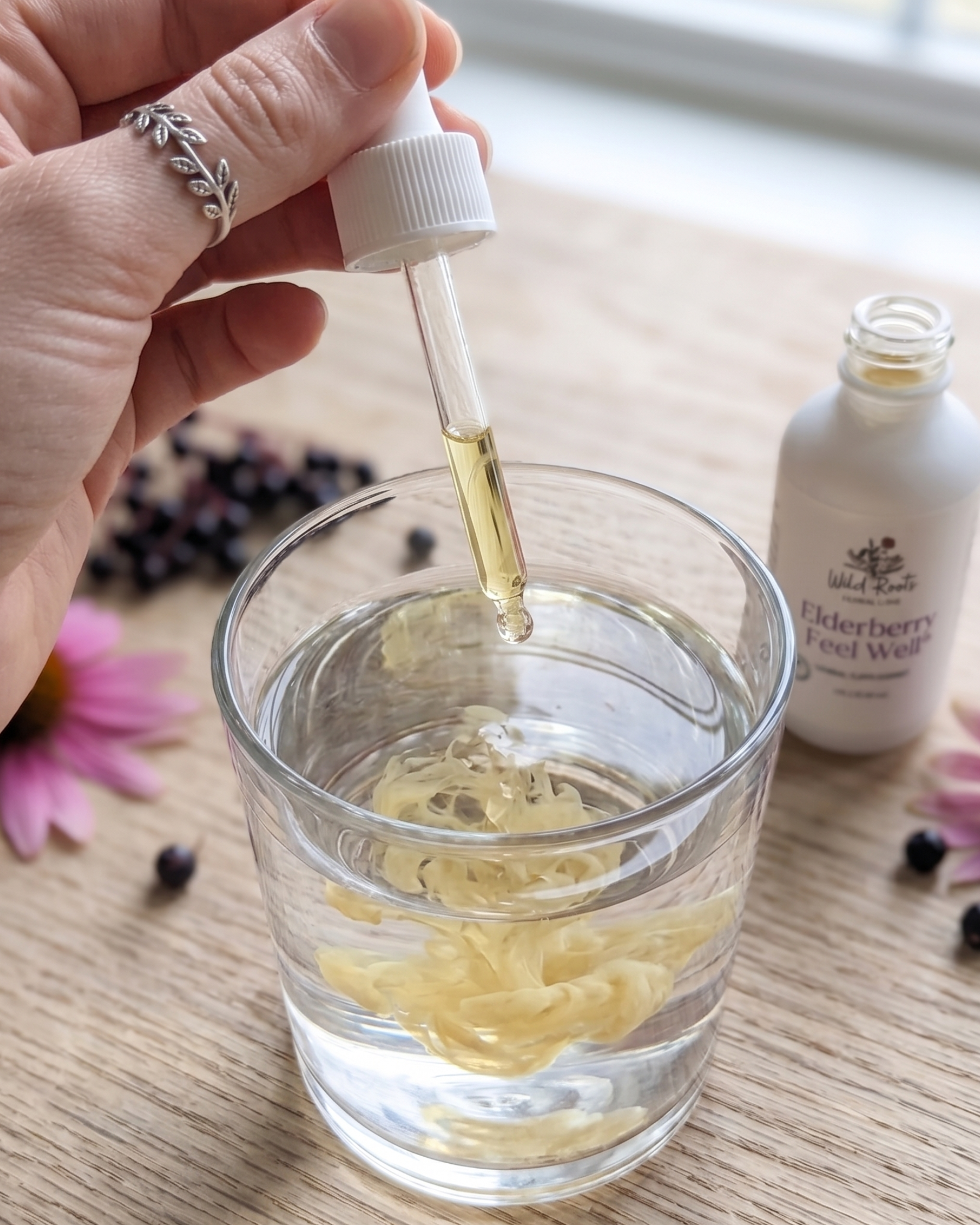 Hand using a dropper to add liquid from a bottle into a glass on a wooden surface with flowers.