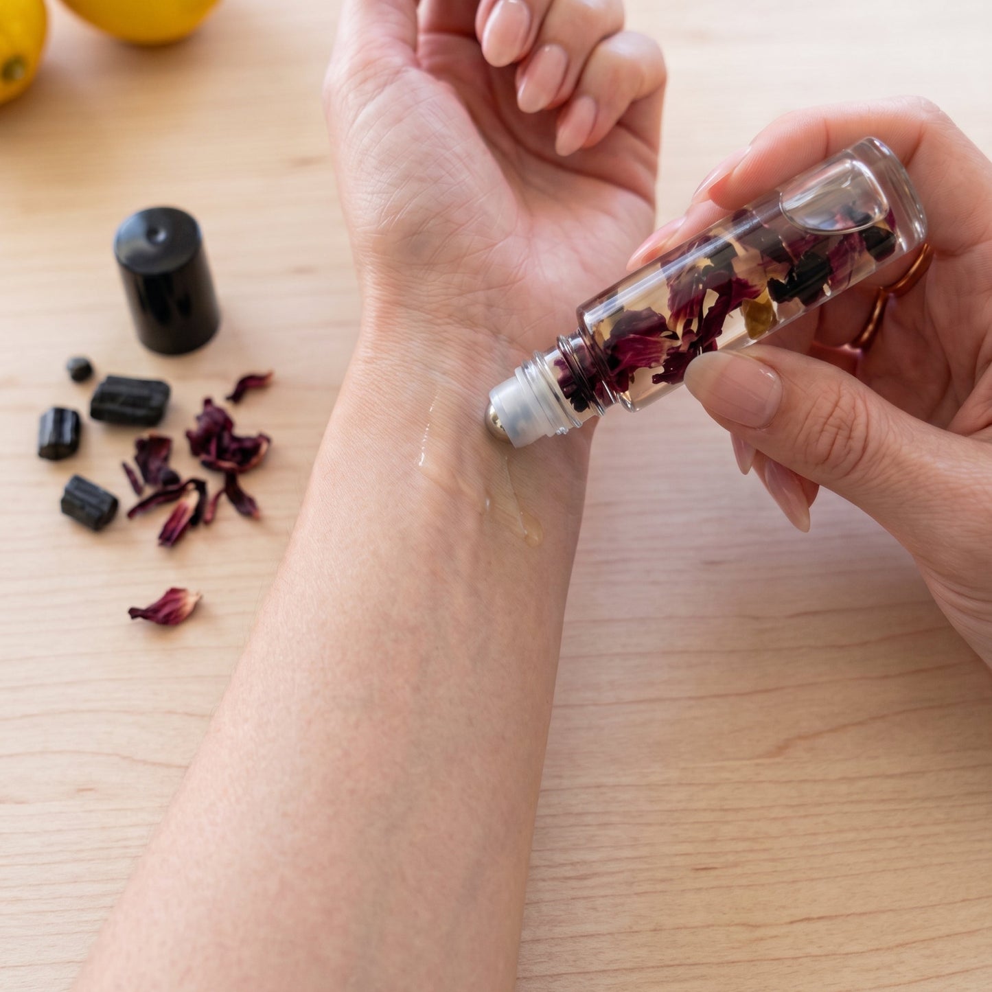 A clear glass roll-on bottle filled with dried flower petals is being used to apply a clear liquid from a clear gel or lotion onto a person's forearm, set on a light wood tabletop with scattered dried petals and a black cap nearby.