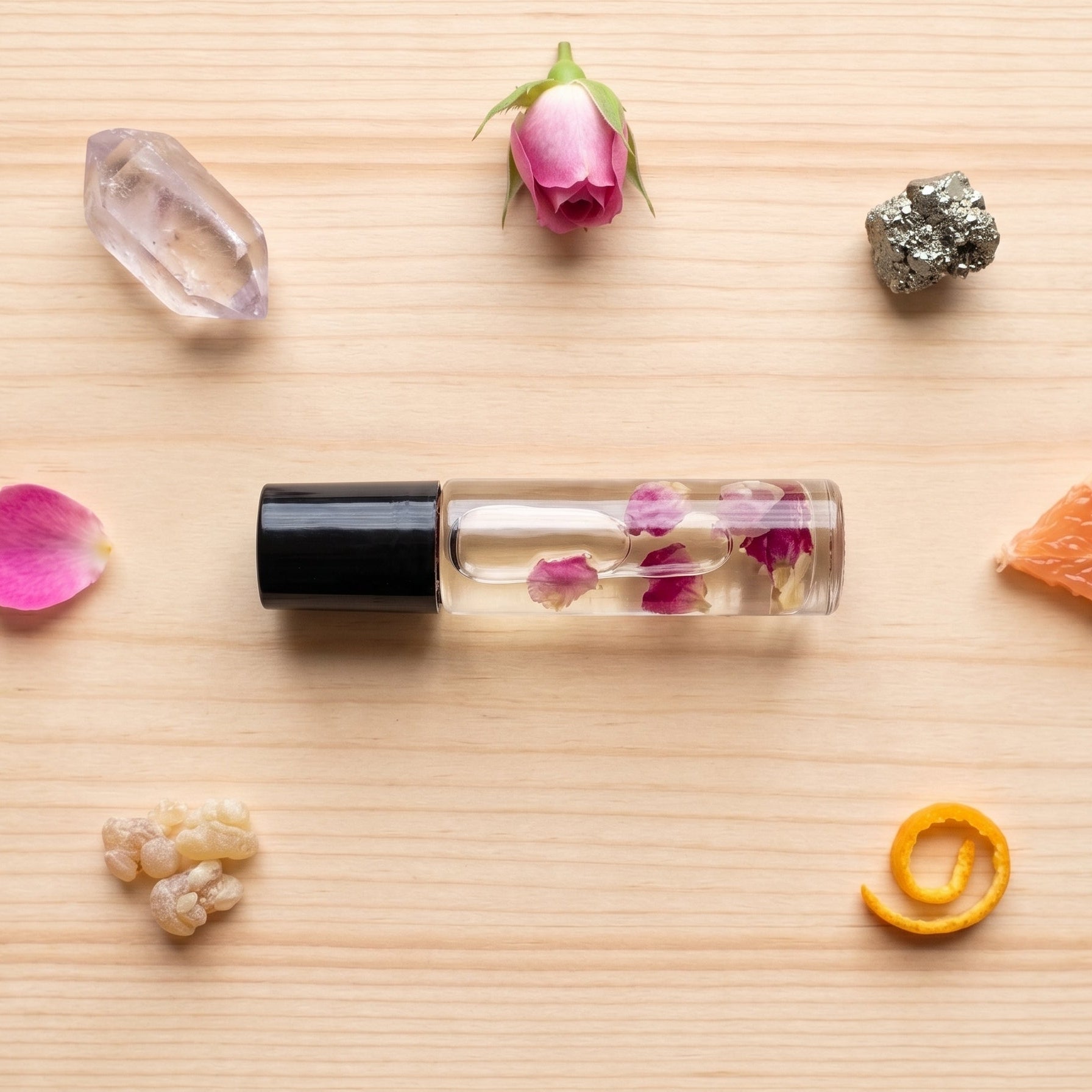A clear glass roller bottle with a black cap, filled with clear liquid and dried pink flower petals, arranged on a light wood surface with botanical and citrus props including a rosebud, crystal, orange slice, and peel.