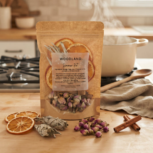 A stand-up pouch of Woodland Scents Summer Fit herbal tea mix stands on a wooden countertop in a warm kitchen setting. The kraft paper bag has a clear window showing dried flowers and orange slices, with steaming tea rising from the package.